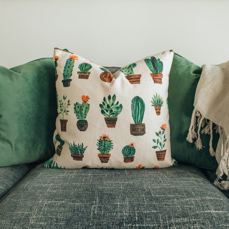 A square, cream-colored throw pillow featuring a grid pattern of various illustrated green potted cacti and succulents sits centered on a textured grey fabric sofa.