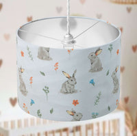 Bunny print kids room drum lampshade in beige, orange, green, blue and white on a blurred background.