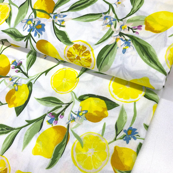 Lemon blossom cotton fabric featuring yellow lemons, citrus slices, blue flowers and green leaves on a white background.