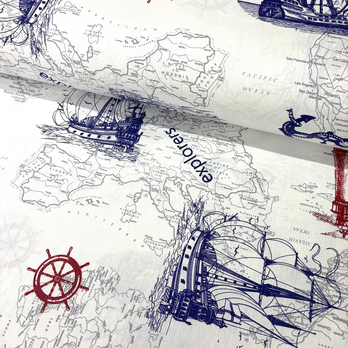 Nautical cotton fabric featuring an antique world map, explorer sailing ships, compass wheel motifs, and marine illustrations in navy blue and red on an off-white background.